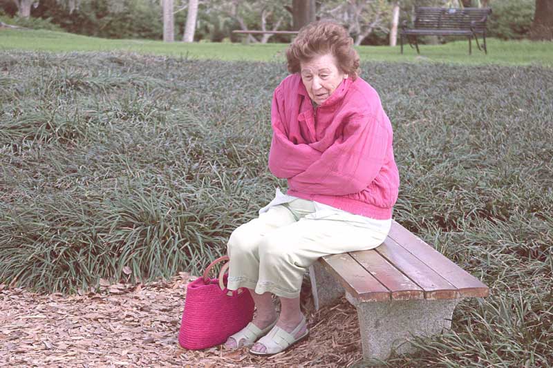 A photo of a senior woman, sitting alone on a bench, looking cold and afraid.